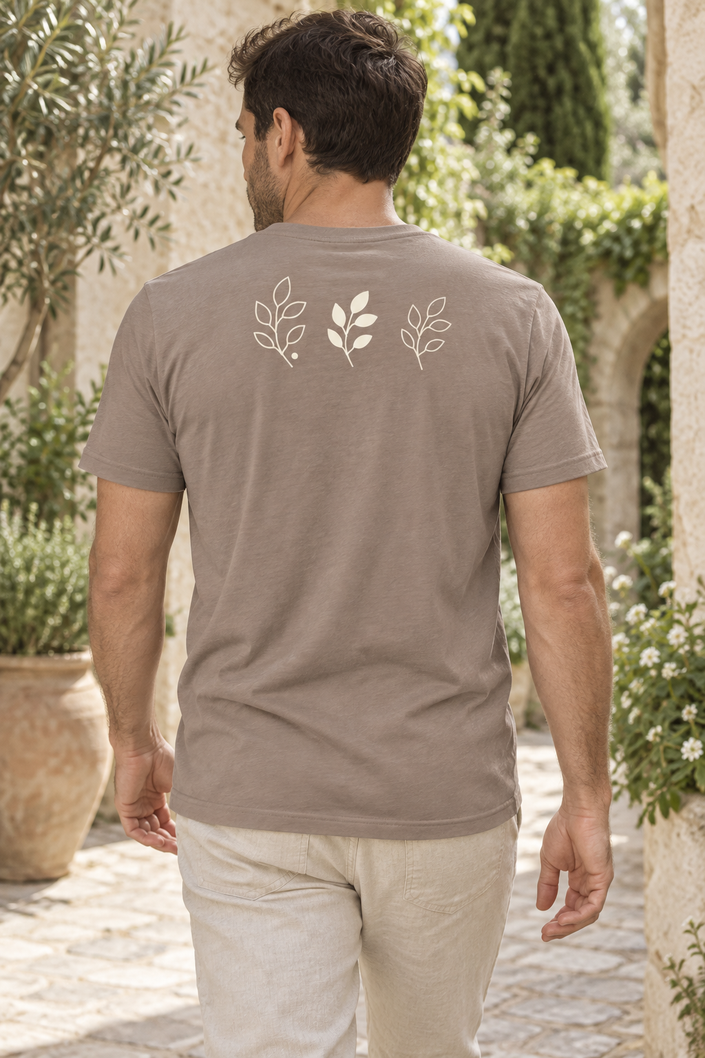 AI image of a fictional adult facing away in a garden courtyard, wearing the Pebble Garden Panel tee with three cream branches across the upper back.